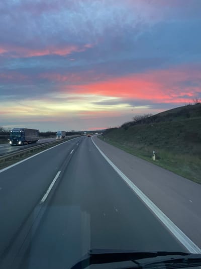 The image depicts a multi-lane highway scene near Wallhausen, Germany, during dusk or sunset. The sky is characterized by prominent bands of red, pink, orange, yellow, and blue clouds. The asphalt highway surface appears wet or reflective. In the direction of travel, a dark-colored passenger vehicle with illuminated headlights is visible in the right lane in the middle distance, with additional vehicle lights further ahead. On the opposing carriageway, separated by a metal guardrail, two dark-colored semi-trailer trucks with illuminated headlights are visible. The right side of the highway features a grassy embankment or hill with sparse vegetation, and a white and black roadside marker post. The foreground reveals the dashboard and a portion of the windshield of the vehicle from which the photograph was taken, with internal reflections visible on the glass.