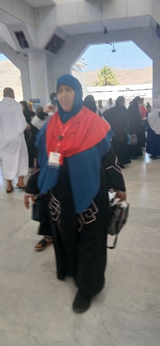 The image captures a bustling public area in Makkah Al Mukarramah, Saudi Arabia, likely an exterior walkway or entrance to a large complex. An adult female, possibly in her 50s-70s, is the central subject, walking towards the viewer, slightly right of the frame's center. She wears a black outer garment (abaya) with light-colored geometric patterns on the sleeves and sides, a blue head covering, and a bright red shawl draped over her shoulders. A red lanyard with a white identification badge hangs around her neck. She carries a dark shoulder bag on her right side and wears dark, closed-toe shoes. Her facial expression is neutral.
To the left and slightly behind the main subject, a man dressed in the two-piece white ihram garment walks away from the camera. Numerous other individuals, including men in ihram and women in various dark clothing (likely abayas and hijabs), are visible in the mid-ground and background, suggesting a gathering of pilgrims. The ground is paved with large, light-colored, possibly marble, tiles.
The environment features substantial light-colored architectural structures, including pillars and an overhead archway. Beyond the archway in the background, a clear blue sky is visible above distant, rugged brown mountains characteristic of the Makkah region. A single palm tree and other sparse green foliage are seen against the mountain backdrop. A building with partially obscured Arabic text on a sign is visible through the arch.