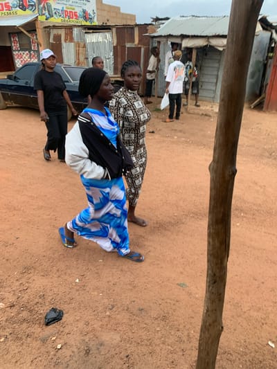 A group of people are gathered on a dusty, unpaved street in Du, Nigeria. A dark blue sedan is parked on the left side of the image. In the foreground, two women are walking towards the right. The woman on the left wears a black and white bomber jacket over a blue and white patterned dress, and blue flip-flops. The woman on the right wears a black headscarf and a black and white patterned dress. Behind them, a man in a white t-shirt and dark pants stands near a makeshift stall with a corrugated metal roof. Other individuals are visible in the background, some standing and some possibly inside the stall. The ground is a reddish-brown dirt and gravel, with scattered debris. The overall scene depicts a bustling street in a Nigerian town.