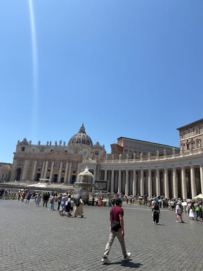 The image depicts a sunny day in Vatican City, focusing on St. Peter's Basilica and the surrounding area. A man in a maroon shirt, khaki pants, and a dark baseball cap is walking across the cobblestone pavement. He is wearing white sneakers and has a black bag slung across his torso. A long queue of people is visible, lined up along a fence in front of the basilica, which suggests a popular attraction. The architecture of the basilica is prominent, with its dome and decorative façade. The sky is a clear blue. The overall scene conveys a sense of tourism in a historic and significant location.