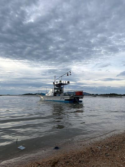 A fishing boat is anchored in the water near the shore. The sky is cloudy, with dark grey clouds covering the upper half of the image. The water is calm with some ripples. The boat is white with a blue stripe along its hull. There is an orange container on the deck of the boat. The shore is made of sand and small pebbles. In the distance, there are islands and the coastline of a city. This scene is located in the city of Ko Samui District, Thailand.