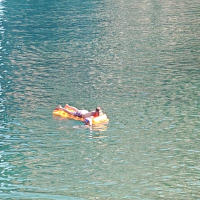 In the turquoise waters of the Black Sea, near the coast of Simeiz, Ukraine, a man and a child are relaxing on an orange inflatable raft. The man, positioned on the raft, is lying on his stomach, wearing purple swim trunks. The child, partially submerged and facing away from the viewer, is positioned in front of the man on the raft. The water's surface is disturbed by ripples, reflecting the sunlight and creating a shimmering effect across the scene. The background consists of the vast expanse of the sea, its color transitioning from a lighter turquoise near the surface to a deeper blue further into the distance. The shoreline or any surrounding land features are not visible in this frame.