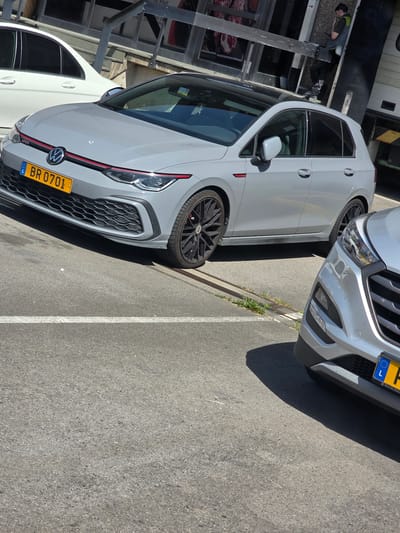 In the city of Leudelange, Luxembourg, a grey Volkswagen Golf GTI is parked on an asphalt surface. The car has a black roof and a license plate that reads "BR 0701". Another car, a silver SUV, is partially visible in the foreground. The background shows a building with a metal railing and a person standing near a loading dock. The scene appears to be a commercial area, possibly a parking lot or a street.