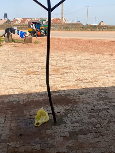 This is a full, eye-level, outdoor shot taken in Kwang, Nigeria, with a focus on a paved area in the foreground and a road and buildings in the background. A black metal pole, part of a structure, runs vertically through the right side of the frame, casting a shadow on the textured, light brown and gray pavement. A crumpled yellow plastic bag lies on the pavement in the lower left quadrant, near a dark, wet patch and some dark debris.

In the mid-ground, a three-wheeled vehicle, commonly known as an auto-rickshaw or "keke napep," is parked. It is green and yellow with the license plate "PTA 9388" visible on the side. Two men are interacting with the vehicle. One, wearing a blue shirt and a pink headwrap, is leaning into the driver's side, holding a red bucket. Another man, in a blue shirt, is partially visible in the driver's seat. A third person, wearing a white shirt, is in the passenger seat. To the left of the vehicle, several pieces of clothing, including black pants and a blue and white shirt, are draped over a concrete block.

The background shows a dusty road, a low brick wall, and several buildings with corrugated metal roofs. Electrical poles and wires are visible against a hazy, pale blue sky. The environment appears to be a somewhat arid, urban or semi-urban setting.