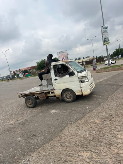 In Karum, Nigeria, on an overcast day, three Black males are traveling in a severely damaged white flatbed truck. The truck's front end is heavily dented and missing its headlight and grille. Two men are seated on top of a load of concrete blocks in the back of the truck, facing forward. One man is leaning back, while the other is seated upright. A third man is in the driver's seat. The truck is on a paved road with some gravel and dirt. In the background, there are buildings, trees, streetlights, and a billboard. A woman in a grey garment and red shoes walks on the side of the road, carrying a load on her head. Another white car is visible in the distance.