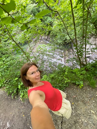 A woman with shoulder-length brown hair, wearing a red sleeveless top and a cream-colored skirt, is taking a selfie while standing on a dirt path. Behind her, lush green trees and foliage are abundant, with a rocky cliff face visible in the background. A waterfall cascades down the cliff, feeding into a pool of water. The location is identified as Izmaylovka, Russia.