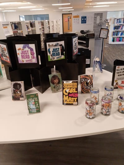 The image presents a well-lit, modern library interior, likely in Coventry, United Kingdom, with a focus on a white, round display table. In the foreground, several black cardboard book stands promote reading by genre, notably "JUST READ ROMANCE" and "JUST READ FANTASY".
Displayed prominently on the table are individual books: "Lionheart Girl", "How To... Save it" (a guide to "FIX YOUR FINANCES"), "Wilder Girls", "Dungeon Crawler Carl" by Matt Dinniman, and "Death an Author" by Nnedi Okorafor. An interactive element is present with multiple glass jars labeled for different genres, including "HEALTH & WELL", "CLASSICS &", "ROMANCE &", "CRIME & TH", and "FA" (likely Fantasy). These jars, filled with small folded papers, are part of a feedback campaign, as indicated by a sign stating, "TELL US WHAT YOU'D LIKE TO READ NEXT" and instructing users to "Put a star in the type of books".
In the mid-ground, a large wall display categorizes books and themes with text such as "RELAX AND RECHARGE", "TALES AND ADVENTURES", "BOOKS", "STORIES", "NOVELS", and "FICTION". A "LOAN POLICY" sign is also visible near a bookshelf. A large printer or copier machine is situated below this display. The background reveals rows of white bookshelves stocked with books, contributing to the bright and organized atmosphere. Modern fluorescent lighting illuminates the space. No people are visible, lending a quiet and inviting ambiance to the scene, suggesting a space designed for reading promotion and community engagement within the library.