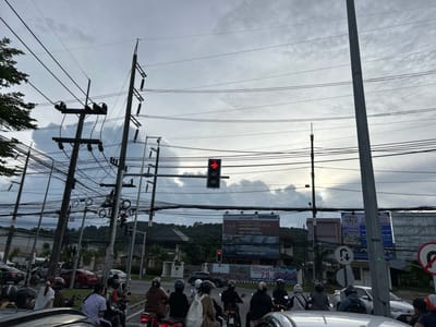 This image captures a street scene in Wichit, Thailand, likely during a cloudy afternoon. The foreground is dominated by a traffic jam, with numerous motorcycles and cars stopped at an intersection. Several people are seen on motorcycles, some wearing helmets and casual clothing.

Above the traffic, a complex network of utility poles and overhead wires crisscrosses the sky, a common sight in many Southeast Asian urban areas. A traffic light prominently displays a red arrow pointing to the right, indicating that traffic from that direction must stop.

In the background, several large billboards are visible, advertising real estate and other services. Some text on the billboards is legible, with Thai script and numbers. One billboard mentions "LAKESIDE POOL VILLA" and a phone number. Another seems to advertise "METAL SHEET" products. Behind the billboards, residential buildings and lush green hills suggest a semi-urban or suburban setting. The overall atmosphere appears to be one of everyday life in a busy Thai town, with the cloudy sky suggesting the possibility of rain or overcast weather.