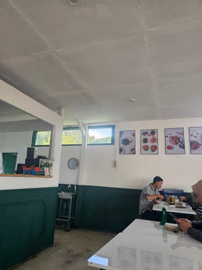 The image shows the interior of a restaurant or cafe. The walls are painted white with a dark green wainscoting. There are framed pictures of food on the wall. In the background, there are windows looking out onto trees and sky. In the foreground, there are several tables. At one table, a man in a grey polo shirt is eating, and a woman wearing a hijab is also seated at the table. Another table in the foreground is empty. There is a sink in the corner of the room. The ceiling is exposed and appears to be concrete. The floor is concrete. The overall atmosphere appears to be casual. The location is identified as Banyuurip, Indonesia, though this specific detail cannot be visually verified from the image alone.