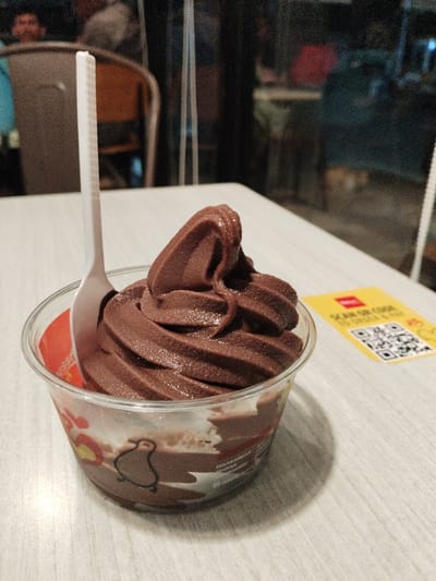 A single serving of chocolate soft serve ice cream in a clear plastic cup with a red and white paper liner is sitting on a light wood-grain table. The ice cream is in a swirl shape, with the top portion of the swirl exposed. A white plastic spoon lies on top of the cup, partially submerged in the ice cream. A small yellow paper card with black text is located to the right of the cup, the text is illegible. The ice cream cup is situated in the foreground of the image. The background shows a blurry scene, likely a cafe or restaurant, with people in the distance and wood chairs and a table to the left and right of the image. The location of the photo is a cafe in the city of Bekasi Regency, Indonesia. The coordinates of the location are -6.298 latitude and 107.149 longitude.