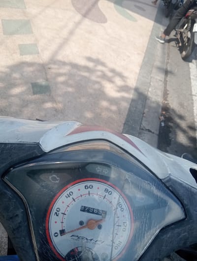 The image shows a close-up of a motorcycle's instrument cluster, with a speedometer displaying a reading of "37963". The speedometer has markings up to 140 km/h. There are also indicator lights for the engine, high beam, and turn signals. The instrument cluster is made of plastic and appears to be slightly worn and dirty.

In the background, a paved walkway with a patterned design is visible, along with a concrete curb and a road. Shadows from trees or other objects are cast on the walkway. A person's legs and feet, wearing dark pants and white sneakers, are partially visible on the right side of the image, suggesting they are either riding or have just dismounted the motorcycle. Another motorcycle is partially visible in the far background.

The overall scene suggests an outdoor environment in an urban or suburban area. The presence of a motorcycle and a paved walkway is common in cities. While the image itself does not explicitly state the location, the request specifies Sidoarjo, East Java.