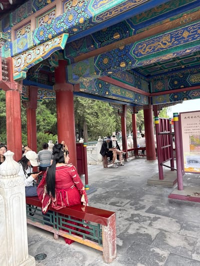 The image depicts a covered outdoor area with ornate, colorful architecture, likely a pavilion or walkway in a traditional Chinese garden. The ceiling is elaborately decorated with blue, green, and gold motifs, including what appear to be dragons and floral patterns. Large red pillars support the roof. In the foreground, a person dressed in a traditional red Chinese outfit with elaborate hair ornaments is seated on a red bench, viewed from the back. To their left, another individual wearing a white cap is also seated and looking at a phone. Several other people are visible in the background, some sitting on benches and others standing, amidst a backdrop of trees. A sign with Chinese characters and imagery is mounted on a purple structure to the right. The ground is paved with stone tiles, some of which appear wet. The overall scene suggests a cultural or historical site within Xicheng District, China, possibly a tourist attraction given the attire of one individual and the presence of visitors.