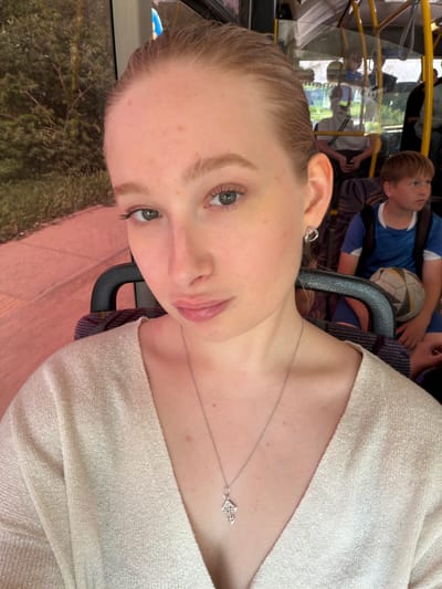 The image shows a young woman on a bus in Rossiyskiy, Russia. She has fair skin, light brown hair, and is wearing a light-colored sweater and a silver necklace with a cross pendant. She is looking directly at the camera. Behind her, through the bus window, trees and foliage are visible. Further back on the bus, a young boy in a blue shirt is sitting with a soccer ball. Other passengers are visible but blurry. A black handrail is near the woman.