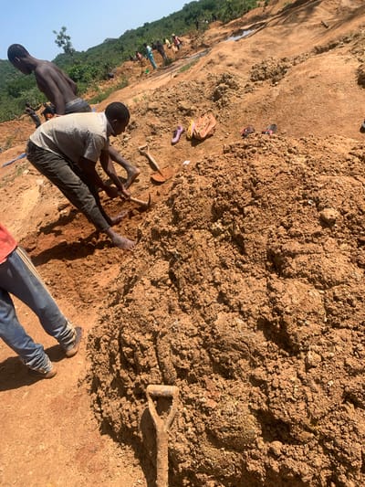 In Buga da Gindi, Nigeria, men work on a construction site on a sunny day. The scene shows a hillside with reddish-brown soil, indicative of the local geology. Several individuals are actively engaged in manual labor, primarily digging and moving earth.

In the foreground, a man wearing a striped shirt and dark pants is crouched, using a pickaxe to break up the soil. His bare feet are visible, stained with dirt. Next to him, a shovel rests upright in a pile of excavated dirt. To the left of the frame, another person's legs are visible, clad in blue jeans and shoes, standing near a pile of excavated earth. In the mid-ground, a shirtless young man wearing grey shorts is bent over, seemingly observing or assisting with the digging. Further up the hillside, a group of men are scattered, some holding shovels and others appearing to supervise or direct the work. The terrain is uneven and appears to be part of a larger earth-moving project, possibly for infrastructure development. Scattered on the ground are personal items, including a brown bag and a pair of flip-flops. The background shows a lush green hillside under a clear blue sky, suggesting a rural or semi-rural setting within Buga da Gindi. The overall impression is one of strenuous physical labor in an outdoor environment.