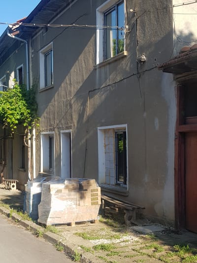 The image depicts the exterior of a two-story building located in the city of Lomets, Bulgaria. The building's facade is made of rough, textured stucco, with visible cracks in places, suggesting an older structure that may be undergoing renovation or is in disrepair. The lower part of the building features several windows and doorways, some of which appear to be under construction or unfinished, with exposed framing around one window and stacks of boxed materials, possibly tiles or building supplies, wrapped in plastic and placed on wooden pallets near the entrance. A vine with green leaves grows up the side of the building, partially obscuring a window. A simple wooden bench sits outside the building. Electrical wires are strung across the upper portion of the facade, and a downspout runs along the side. The scene is bathed in sunlight, casting shadows across the building and the surrounding paved area. A street or sidewalk runs along the bottom of the image, with grass and weeds growing along the edge. The overall impression is of a residential or commercial building in a semi-urban or rural setting, likely in a state of work or neglect.