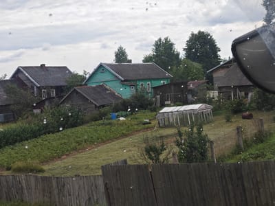 The image depicts a rural landscape in Yamskaya Sloboda, Russia, under a cloudy sky. In the foreground, a weathered wooden fence runs horizontally across the frame, with visible grain and some sections appearing aged. Beyond the fence, a sloping grassy area is visible, dotted with small white specks, which could be water droplets or debris. Further up the slope, rows of green plants, possibly a garden or crops, are arranged in neat lines.
Several structures are present in the midground. A greenhouse with a glass roof and metal frame sits on the right side of the slope. To its left, a collection of buildings forms a village-like setting. These include a two-story green house with a dark roof, several wooden outbuildings with corrugated metal roofs, and a larger wooden structure with a dark, shingled roof and a chimney. Trees and dense green foliage are interspersed throughout the scene, with some appearing to be fruit trees or bushes.
In the background, more trees and buildings are visible, suggesting a larger settlement or a densely wooded area. The overall impression is one of a quiet, rural setting, possibly a dacha or village in the Russian countryside. The image has a slightly blurry quality, possibly due to moisture on the lens or the camera used.