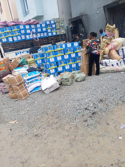The image captures an outdoor scene in Satellite Town, Nigeria, depicting the loading or unloading of a delivery truck. The ground is a mixture of grey gravel and reddish-brown dirt.

In the foreground, various goods are stacked. On the left, several bundles wrapped in brown paper and secured with white straps are piled. To their right are multiple white sacks, some with blue branding, lying on the ground. Next to these are smaller, green-wrapped bundles. A large white sack, possibly containing textiles, bears the partially visible text "CHID[...]" on its side.

Towards the center and right, a large white truck is backed up against a grey building. The truck's side has the visible word "PICK" in blue lettering. The truck bed is laden with numerous blue boxes, uniformly stacked. These boxes feature a camera or lens icon, along with text including "SUPERIOR QUALITY" and "QUALITY GUARANTEED" in English, and some Arabic script. Some red and black coiled items, possibly ropes or hoses, are also visible on top of the stacked boxes in the truck.

Several people are present, engaged in handling the goods. In the truck bed, a man in a dark t-shirt is seen holding a coiled red and black item, appearing to be either loading or unloading. Another man in a dark shirt is partially visible further inside the truck. Standing to the right of the truck, facing away from the camera, is a woman wearing a colorful patterned top and dark trousers. She is positioned near a stack of cardboard boxes, some of which are marked "FRAGILE." Behind her, piles of large beige sacks are stacked against the building. In the background, near the building's entrance, two other men are visible, one in a blue shirt.

The scene appears to be bustling with commercial activity, likely a goods transfer point. The lighting suggests it is daytime with bright, clear weather.