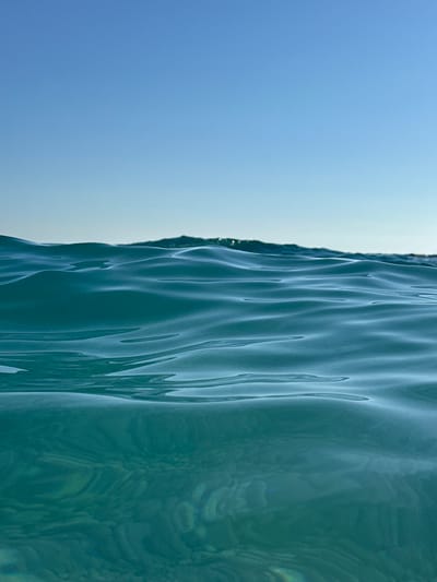 The image displays an abstract close-up of a seascape, characterized by a distinct division between the sky and the water, possibly taken from a very low angle at the waterline or partially submerged. The left side of the frame features a clear, bright blue sky, gradually lightening in hue towards the center, suggesting a clear day. On the right, the turbulent surface of the sea is visible, rendered in shades of teal and turquoise.

The water exhibits numerous small, distinct ripples and undulations, which catch and reflect sunlight, creating dynamic vertical patterns and subtle swirling textures across its surface. A sharp, irregular line separates the tranquil expanse of the sky from the active, reflective water. The perspective emphasizes the intricate details of the water's surface and its interplay with light. The scene conveys a sense of natural beauty and serenity. No activity or event is explicitly depicted beyond the natural movement of the water. There is no visible text in the image. Given the context, the location is likely the Mediterranean or Aegean Sea near Kaş, Turkey, under fair weather conditions during the daytime.
