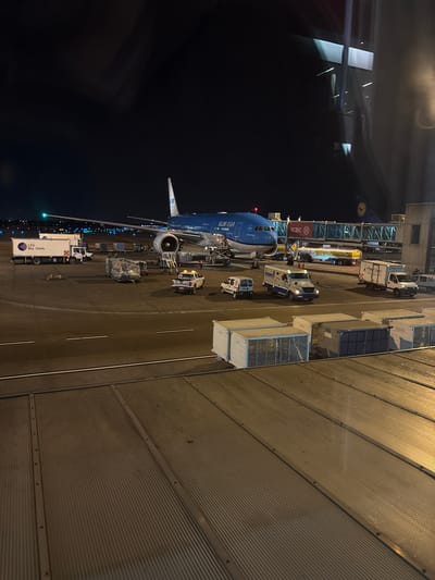 The image shows a nighttime scene at an airport, likely Aeropuerto Internacional Ezeiza, Argentina, with a KLM airplane parked at a gate. The airplane is a prominent subject, its blue and white livery visible, with "KLM" clearly legible on its side. Various ground support vehicles surround the aircraft, including a catering truck with "Sky Chefs" printed on it, a baggage loader, and other utility vehicles. A jet bridge connects the terminal building to the plane. The terminal appears to be a modern structure with glass and metal components. The overall setting is an airport tarmac at night, illuminated by runway lights, vehicle headlights, and the lights from the terminal. The ground is paved with asphalt or concrete, and ribbed metal panels line the side of the image, possibly part of the terminal building or a service area. The scene depicts the operational activities at an airport after dark, with ground crews likely preparing the aircraft for its next flight or servicing passengers. There is no visible indication of weather conditions, but the darkness suggests it is nighttime. No specific emotions are conveyed, and the activity is purely functional and operational. Visible text includes "KLM", "Sky Chefs", and "ICBC".