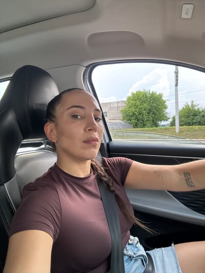 A woman is sitting in the driver's seat of a car, looking at the camera. She has her hair in a braid and is wearing a maroon t-shirt and denim shorts. She has a tattoo on her left arm. The car appears to be in motion, as indicated by the scenery outside the window, which includes trees and buildings. The scene takes place in the city of Krasnaya Pakhra, Russia.