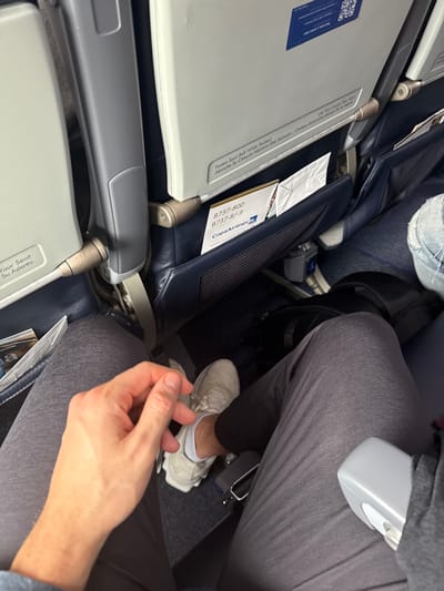 An interior shot captures the confines of airplane seating. Two rows are evident, with folded-down tray tables featuring safety instructions and seat information in both English and Spanish. A Copa Airlines brochure is visible in the seat pocket of the row in front. Passengers occupy the seats, their legs and feet encroaching on the limited space. One person is wearing light-colored sneakers with white socks. The overall scene conveys the typical cramped conditions of air travel.