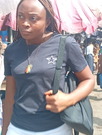 The image captures an outdoor market scene in Asaba, Nigeria. A female individual with brown skin and braided hair, wearing a dark blue short-sleeved t-shirt featuring a white star outline logo on the left chest and white lower garments, is prominently featured. She wears a necklace with a multi-colored rectangular pendant and carries a dark-colored shoulder bag on her right side, held by her right hand. Her gaze is directed towards the left of the frame. The background depicts a bustling market environment with blurred figures, multiple reddish-brown market umbrellas, and an unpaved ground surface, illuminated by bright daylight.