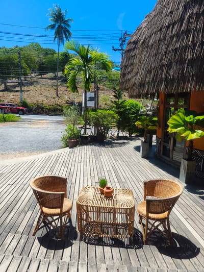 This is a high-angle, sunlit outdoor photograph taken at what appears to be a resort or cafe in Baan Thong Po, Thailand. In the foreground, a wooden deck is furnished with two woven rattan chairs and a small, similarly constructed table. The table holds two small terra cotta pots, one with a sprig of greenery. The deck planks are weathered and laid horizontally. Beyond the deck, a gravel area and a road are visible, with a red vehicle parked on the side. A lush, green hillside with trees, including a tall coconut palm and a smaller fan palm, rises in the background. Overhead, power lines crisscross the bright blue sky. To the right, a building with an orange exterior features a thatched roof and large windows. The overall scene suggests a tropical, relaxed atmosphere.