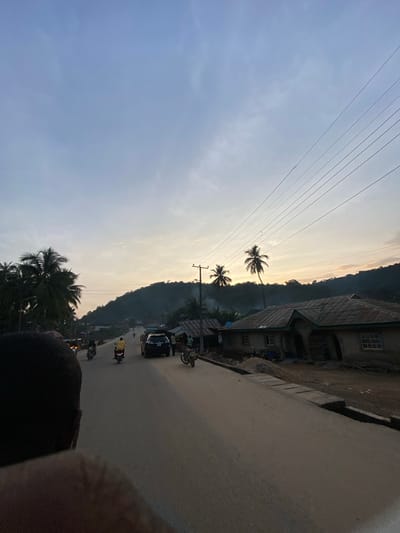 This image captures a street scene in Igarra, Nigeria, during either dawn or dusk, indicated by the warm, low-angle light on the horizon blending with a blue-grey sky. The sky occupies the upper two-thirds of the frame, transitioning from a pale golden hue near the horizon to a desaturated blue-grey overhead, with wispy clouds visible throughout. Multiple power lines traverse the upper right quadrant of the sky, slanting downwards to the right.

In the midground, a paved road extends from the foreground into the distance, ascending slightly towards a prominent, tree-covered mountain range that dominates the background. The mountains are silhouetted against the lighter sky, appearing dark green to black. On the left side of the road, dense tropical foliage, including several palm trees, partially obscures a dark, indistinct shape in the immediate foreground, likely a person's head and shoulder, suggesting a view from a moving vehicle.

Vehicular traffic on the road includes at least two motorcycles, each carrying a single rider, one in a yellow shirt, moving away from the viewer. A dark-colored SUV is parked or moving slowly on the right side of the road, further down. On both sides of the road, structures consistent with residential or small commercial buildings are visible. On the right, a single-story building with a multi-pitched, corrugated metal roof and white-framed windows is prominent. It appears to be constructed of earth or cement with some visible wear. Next to it, another smaller, simpler structure is visible. A utility pole stands on the right, supporting power lines. Other smaller structures and vegetation are scattered along the roadside, extending towards the distant base of the mountains. The overall impression is that of a bustling local road within a town nestled amidst a natural, hilly landscape.