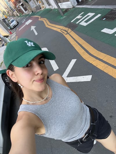 A young woman with dark hair, wearing a green baseball cap, a gray tank top, and black shorts, poses for a selfie on a street in Daegu, South Korea. She has a pearl necklace with a small silver charm and pearl earrings. The street is marked with yellow and white lines, and green painted sections with Korean characters and arrows. A silver car is parked on the left side of the street, and buildings line the background.