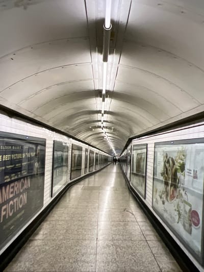 The image shows a long, arched subway tunnel with white tiled walls and a light-colored floor. Fluorescent lights are mounted along the ceiling. On the left side of the tunnel, there are large posters displayed behind glass, one of which advertises the movie "American Fiction." On the right side, there are also posters displayed behind glass, featuring images of food. The tunnel recedes into the distance, with two figures visible walking away from the camera. The environment appears to be a public transportation hub, likely a subway station in the city of Verona, Italy, given the context.