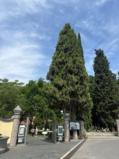 The image shows the entrance to a park in Sirmione, Italy, with two people standing near the gate. The park features tall cypress trees and lush greenery, with a path leading into the park. The sky is blue with scattered clouds. The entrance is marked by stone pillars and metal gates, with informational signs posted. The overall scene is one of a well-maintained public space on a sunny day.