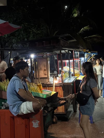 The image depicts a night scene at a street food market in Canggu, Indonesia. A woman with long dark hair, wearing a gray shirt and denim shorts, sits on a small blue stool, using a long metal utensil to stir something in a wok. Several people stand behind her. In the foreground, a man wearing a light blue t-shirt is sitting and smoking a cigarette. He is surrounded by ears of corn. The background features food stalls brightly lit with neon lights, displaying various items for sale. Tropical foliage is visible in the periphery of the scene.