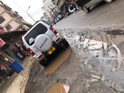 The scene is located in the city of Arusha, Tanzania, at the coordinates -3.372 latitude and 36.688 longitude. A silver Suzuki Sport SUV is parked in a street with a significant amount of mud and potholes. The vehicle is facing towards the right side of the image, and its rear is visible, including a spare tire and a license plate. There is another vehicle parked in the distance, and several people are visible walking in the street, but they are blurred. The background consists of buildings with visible windows and a power line. The street is covered in mud and water, with several puddles present. The scene suggests a recent rainfall or a general condition of poor infrastructure in the area.