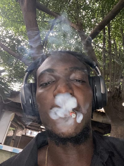 A close-up, rotated image shows a dark-skinned male wearing headphones, exhaling smoke. He has a beard. Behind the smoke and the man are trees and buildings. The location is possibly Alagba, Nigeria.