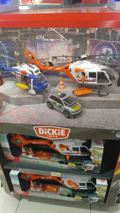 The image shows a retail display of toy rescue vehicles. In the foreground, a display box for "Dickie Toys" rescue helicopters is visible. Two toy rescue helicopters are partially visible inside the box, along with orange traffic cones and a stretcher. Above this box, a diorama features toy rescue vehicles, including a blue and yellow rescue helicopter, a white and orange rescue helicopter with a basket hanging from it, and a silver police car. The backdrop of the diorama is a colorful mural depicting a cityscape with a Ferris wheel and palm trees. The overall scene suggests a toy store or a similar retail environment. The provided crops do not offer additional context about the location within the city of Pylaia Municipal Unit, Greece.