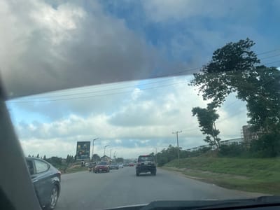 The image shows a view from inside a vehicle, looking out at a road with traffic. The scene is set in Galadimawa Village, Nigeria. It appears to be daytime, with a cloudy sky that suggests it might be overcast or that rain is possible.
Several cars are visible on the road, indicating moderate traffic. In the foreground, a dark-colored car is partially visible on the left. Further ahead, there are more cars, including a red SUV and a black SUV. On the right side of the road, there is a grassy embankment with trees and some construction or new buildings in the background.
A prominent billboard is visible on the left side of the road, advertising "ORIJIN" with the text "NOW IN 33CL". Telephone or power lines stretch across the sky, supported by utility poles. The overall mood of the image is neutral, capturing a typical scene of road travel. The clouds suggest a diffused light, rather than bright sunshine.