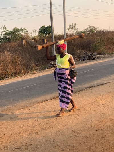A woman in a yellow top, patterned skirt, and red hat carries a large wooden object on her shoulders while walking along a road. A small child is strapped to her back. She wears flip-flops. The road is paved on one side and dirt on the other. Vegetation, including trees and brush, borders the road. Utility poles are visible in the background.