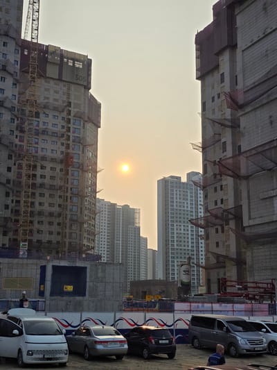 The image depicts a construction site in Daejeon, South Korea, likely a residential area given the presence of numerous apartment buildings in various stages of completion. The foreground features several parked cars, including a white Hyundai Staria, a silver sedan, and a gray minivan. A construction worker in a white hard hat and reflective vest is visible standing near a concrete barrier. The middle ground shows more of the construction site with concrete structures, barriers, and what appears to be a concrete silo with "ASIA" branding. In the background, several tall apartment buildings are visible, with some under construction and others appearing complete. The sun is setting or rising, casting a warm glow over the scene, and the sky is hazy. The overall impression is one of ongoing urban development in a modern city.