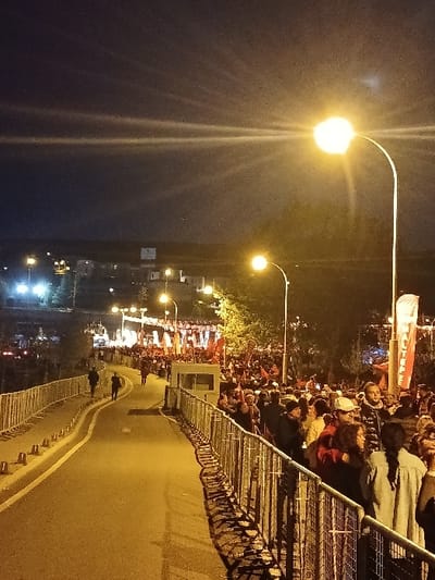 The image captures a nighttime scene in Istanbul, Türkiye, at the coordinates 41.083 latitude and 28.677 longitude. A large crowd of people is gathered behind a metal fence along a paved path that winds through a park. Several streetlights illuminate the scene, casting a warm glow on the crowd. Some people are wearing hats and jackets. There are signs and banners in the background. The street is lined with trees on either side. The sky is dark and clear. The camera is positioned at the end of the path looking down at the crowd. There are two figures walking on the path, one is walking toward the camera and the other is walking away. There is a security booth in the middle ground. The image was taken at night.