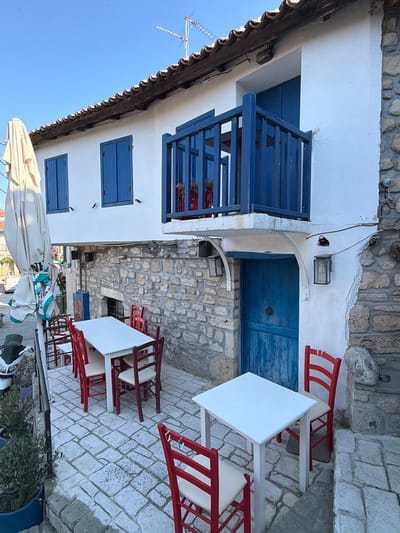 A charming outdoor seating area of a Greek taverna is depicted in this image, likely in Kassandra Municipal Unit, Greece. The scene is bathed in bright sunlight, casting sharp shadows. A white, canvas-covered umbrella stands to the left, partially obscuring a white scooter parked nearby. In the foreground, several white square tables are set with red-trimmed chairs on a cobblestone patio. A larger rectangular table, also white with red chairs, is situated further back. The building itself features a white stucco facade adorned with blue shutters and a blue wooden balcony with a matching railing. A large blue wooden door is visible below the balcony, set into a stone-clad lower level. The building's roof is covered with traditional terracotta tiles. The overall impression is one of a tranquil, inviting establishment ready to welcome patrons.