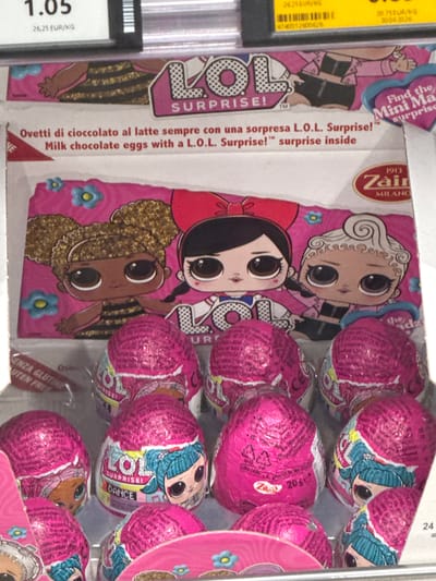 This media file shows a close-up view of a retail display of L.O.L. Surprise! milk chocolate eggs. The foreground is dominated by the bright pink packaging of the chocolate eggs, each individually wrapped in metallic pink foil decorated with images of L.O.L. Surprise! dolls. Some of the text on the packaging is visible, including "LOL SURPRISE!", "DANCE", and Italian text that translates to "Milk chocolate eggs with a L.O.L. Surprise! surprise inside".

Behind the eggs, a larger cardboard display featuring illustrations of L.O.L. Surprise! dolls is visible. The dolls are depicted in a playful, cartoonish style with large eyes and vibrant outfits. Text on this display includes "Find the Mini Ma" and "SURPRISE!". The brand "Zain MILANO" is also present, suggesting the chocolate manufacturer.

On the left side of the image, a yellow price tag with black text is partially visible, showing a price of "26,25 EUR/KG" and an expiration date of "30.06.2026". This, along with the presence of multiple identical products arranged for sale, indicates a retail or point-of-sale setting. The overall impression is that of a commercial display of a popular children's toy and confectionery item. The lighting is bright, suggesting an indoor, well-lit environment, typical of a shop. There are no people visible in the frame.
