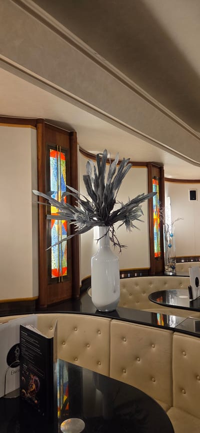 The image depicts the interior of a restaurant or bar, likely in Kehl, Germany, given the metadata. The setting is characterized by a curved booth seating area with light beige, tufted upholstery and dark wood trim. In the foreground, a black, glossy table is visible, holding a menu stand with a black menu featuring an image of fruit. A white, opaque vase containing a large, grey, artificial floral arrangement is centrally placed on the table.
The background reveals architectural details, including wooden panels with stained-glass insets featuring a colorful, geometric design. The ceiling features ornate molding. To the right, another table with similar seating is partially visible, as well as a tall glass vase containing decorative branches and blue spheres. The overall lighting is soft and ambient, creating a sophisticated atmosphere. No people are present in the image.