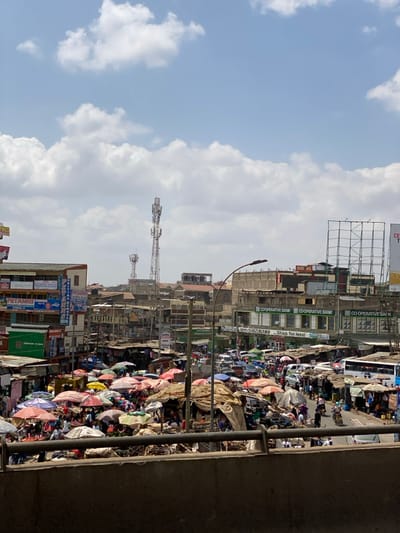 The image shows a busy street market in Githurai, Kenya, taken from a high vantage point. The view looks down upon a bustling market scene, with numerous vendors selling goods under brightly colored tarpaulins and umbrellas. The image is taken from an elevated location, likely a bridge or walkway, providing a bird's-eye view of the market and surrounding buildings. The foreground is dominated by a cement wall, which partially obscures the market activity. The market extends back to the left, where it meets a series of multi-story buildings. In the background, a cell phone tower is visible against a partly cloudy sky. The buildings on either side of the market display various signs, including those for Cooperative Bank and a shop for sale. There are many people in the market, walking between stalls, haggling with vendors, or carrying goods. The scene is characterized by a sense of organized chaos, with the colors of the umbrellas and merchandise contrasting with the gray concrete of the buildings and the bright blue sky. The overall impression is one of a vibrant and energetic commercial hub.