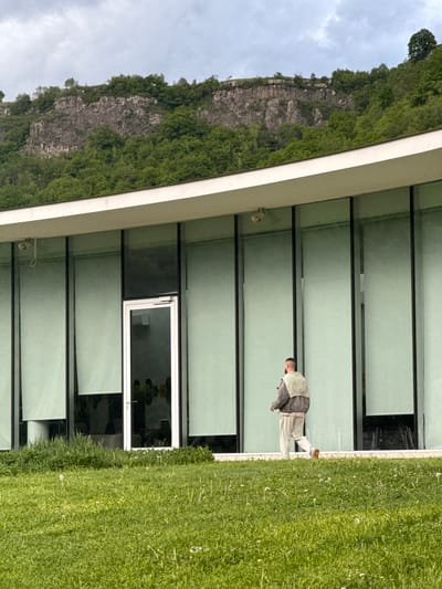 The image shows a man walking on grass in front of a modern building with large windows, located in Chkalov, Armenia. A rocky, green cliff is visible to the left of the building. The sky is overcast. The man is wearing light-colored pants, a light shirt, and a darker jacket. There is a security camera mounted on the building's exterior.