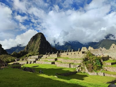 This wide-angle photograph presents a panoramic view of the ancient Inca citadel of Machu Picchu in Peru, showcasing its iconic stone architecture and stunning natural surroundings. The scene is dominated by meticulously constructed stone terraces and the ruins of multi-level stone buildings, which stretch across the midground from left to right. These structures are built with characteristic dry-stacked stone masonry, forming walls and foundations that climb the mountainside.
Lush, vibrant green grass covers the terraced fields and open areas within the ruins. Patches of sunlight illuminate certain sections of the grass and stone, while other areas are cast in shadow, indicating bright, directional lighting, likely from a strong sun on a partially cloudy day.
In the background, a majestic, steep-sided mountain peak, likely Huayna Picchu, rises prominently on the left, covered in dense green vegetation. A range of other verdant mountains extends across the horizon, some of their peaks partially obscured by dense white and gray clouds, giving a sense of the high-altitude, often misty environment. The sky above is a mix of clear blue patches and dramatic white clouds, adding depth and contrast to the landscape.
Several small groups of people, presumably tourists, are visible exploring the site, scattered across the grassy areas and near the stone structures. They appear small against the immense scale of the ruins and the surrounding mountains, emphasizing the grandeur of the location. The overall impression is one of awe-inspiring historical architecture set within a breathtaking natural landscape, bathed in the light of a dynamic sky. No discernible text is visible in the image.