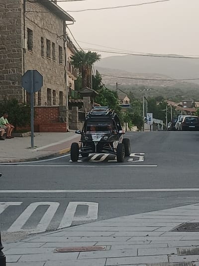 In Navaluenga, Spain, on an overcast day, a dark-colored, open-top buggy with a roof rack drives over a speed bump on an asphalt road. The buggy has large, off-road tires and a visible roll cage. In the foreground, the word "STOP" is painted in large white letters on the road surface. To the left, a man in a light green t-shirt and shorts sits on a low brick wall near a building with a stone facade and multiple windows. Behind the buggy, several cars are parked on the right side of the road, and a tall, thin transmission tower stands further down the street. The background features a hazy, mountainous landscape with trees and scattered buildings.