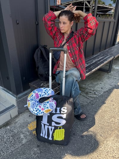 A person with brown hair pulled back into a ponytail sits on a wooden bench, facing right. They are wearing a red and black plaid shirt, a brown t-shirt, and blue jeans. Their right arm is raised and bent, with their hand resting on their head as they adjust their hair. In front of them, on the ground, are a black backpack, a rolling suitcase with a floral-patterned neck pillow on top, and a black bag with white lettering that says "THIS IS MY" and a yellow suitcase graphic. The person is wearing black slide sandals. The scene appears to be an outdoor waiting area, with a dark grey wall to the left and a sunny, textured ground surface in front. The background shows an overhang structure with metal supports and solar panels, suggesting a public transportation stop or outdoor facility. The overall lighting indicates it is daytime. This image was captured on the island of Jeju, South Korea.