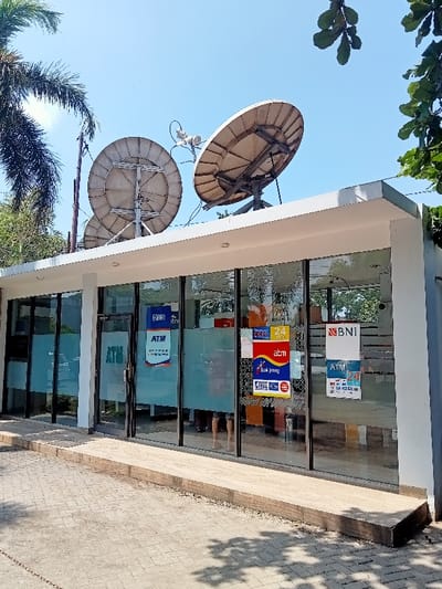 The image shows a white building with large windows, located at coordinates -6.76, 111.015 in Kecamatan Margorejo, Indonesia. Two satellite dishes are mounted on the roof of the building. The building is an ATM kiosk with several signs indicating various banks, including "BNI" and "Bank Jago", with "ATM" printed on them. There are no people visible inside the kiosk, but a person can be seen through the glass door, with the bottom half of their body visible. The front of the kiosk has a step leading up to the entrance. The ground in front of the building is made of cobblestones. There is a palm tree visible on the left side of the image. The sky is a clear blue.