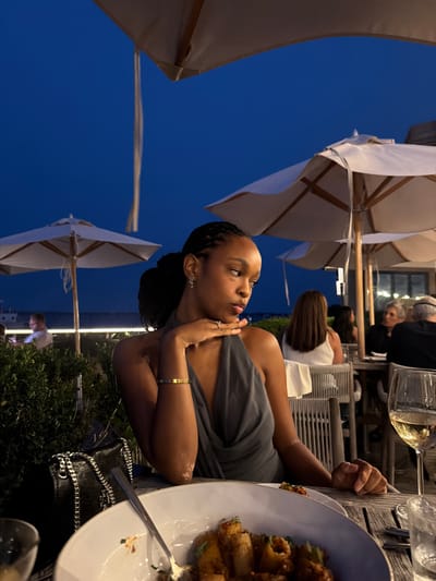 An outdoor dining area at dusk, likely in Montauk, New York, features a young Black woman seated at a table with a plate of pasta and a glass of white wine. She is wearing a grey halter dress and has her hair braided. Her left arm rests on her chin, and she wears a gold bracelet and a ring. In the background, other diners are visible, and umbrellas provide shade. The sky is a deep blue, indicating the setting of the sun.