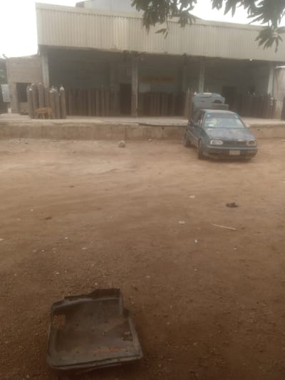 The image shows an outdoor scene in what appears to be a commercial or industrial area. The location is Television, Nigeria. A large, corrugated metal building with an open front dominates the background, with rows of tall, cylindrical gas tanks visible inside. A blue-green car is parked in front of the building, with its trunk open and tires piled inside. A dog is standing to the left of the gas tanks. In the foreground, there's a dirty, rectangular car floor mat. The ground is dirt and scattered with debris. The lighting suggests it is daytime, possibly overcast.